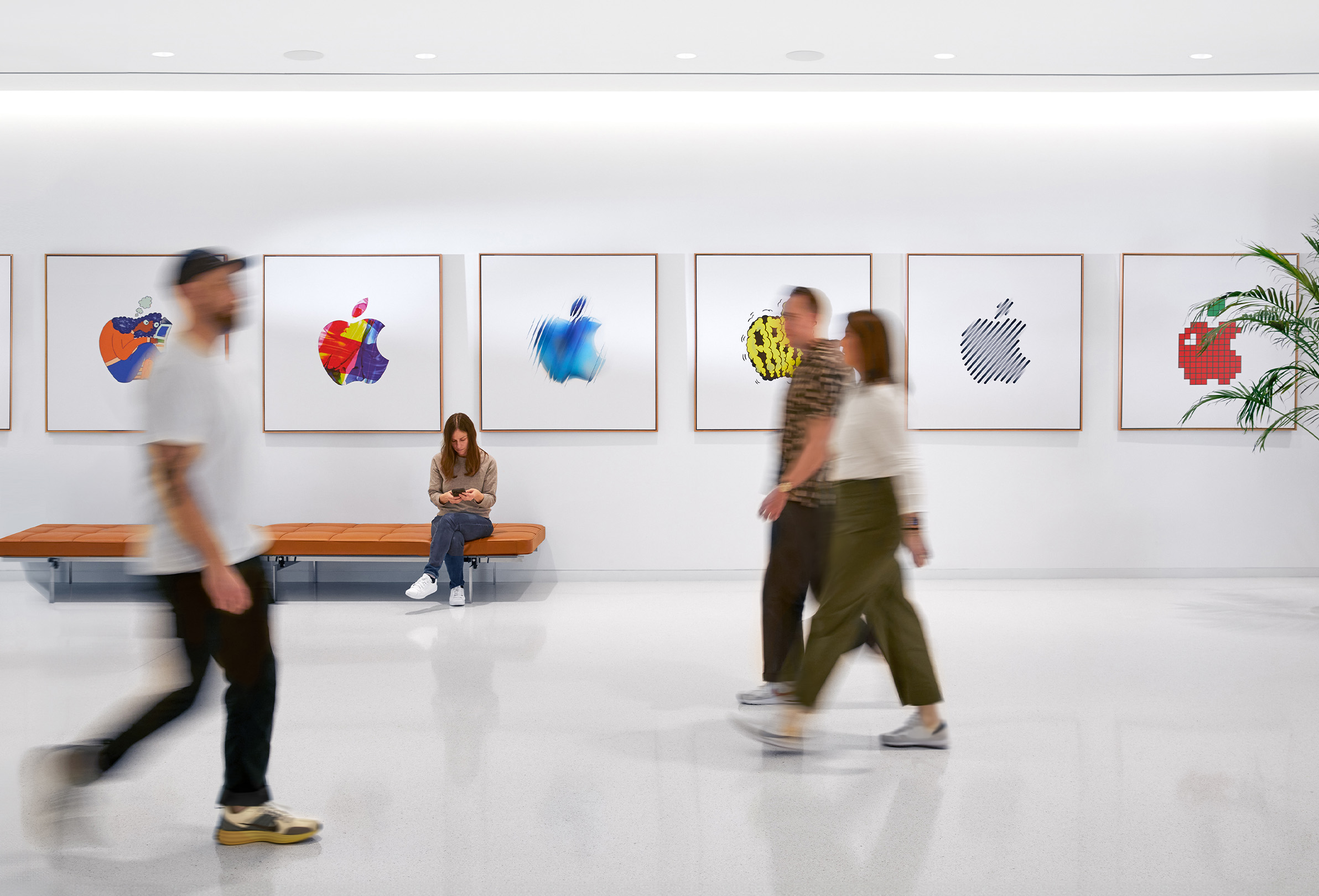 Drei Apple Teammitglieder, die in einem Gebäude vor einer Wand mit farbenfrohen Apple Logos vorbeigehen. Ein viertes Teammitglied sitzt auf einer Bank.
