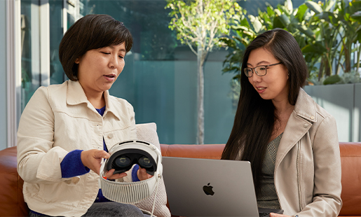 Jessica sitzt in einem Raum an einem MacBook, die Tür zum Innenhof ist offen. Neben ihr sitzt ein Apple Teammitglied, das ihre Apple Vision Pro zeigt.