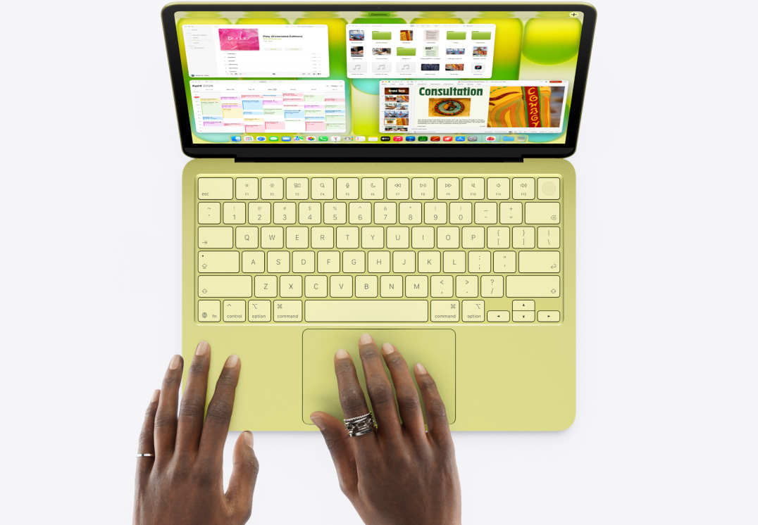 Citrusově žlutý MacBook Neo s něčí rukou na trackpadu, na obrazovce jsou otevřená okna různých aplikací včetně Hudby, Kalendáře a Microsoft PowerPointu.