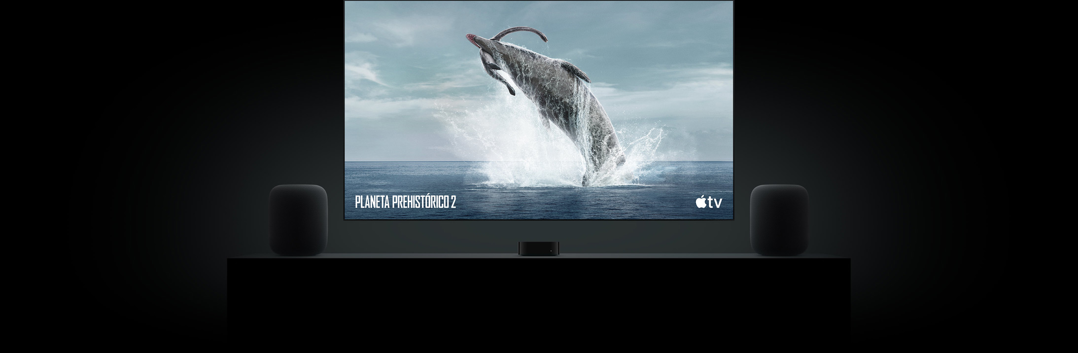 Televisor de gran tamaño con unos dinosaurios de «Planeta prehistórico» a todo color. Está colgado encima de un mueble de televisión con un Apple TV en medio y un altavoz HomePod a cada lado.