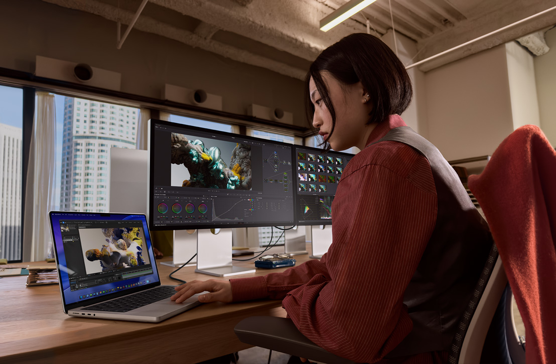 A person works at a desk with a MacBook Pro connected to external display, both screens showing a 3D render, buildings visible through the window behind