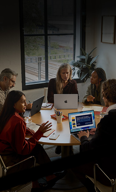 Five individuals seated around a conference table, collaborating on MacBooks, with iPhones face down on the table