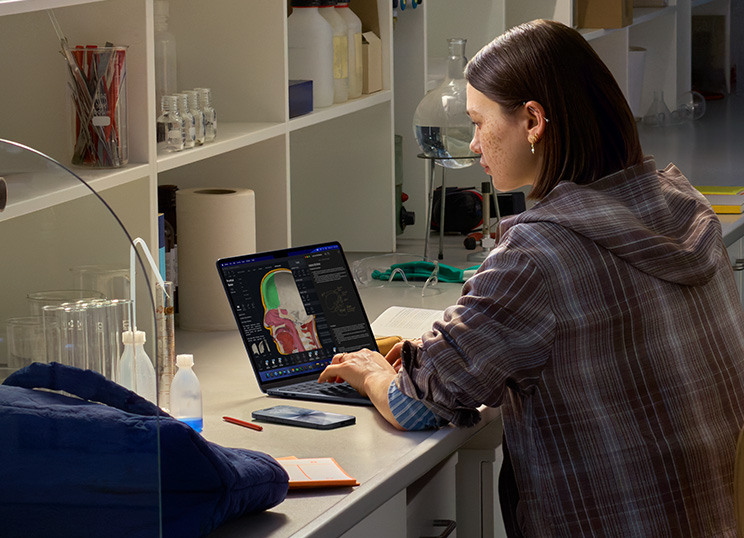 A person sitting in a lab using MacBook, screen displaying multiple apps.