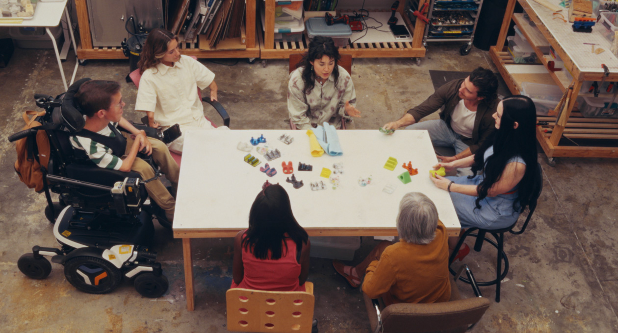 A group of people with different mobility needs sitting together at a table while looking at an array of colorful adaptive smartphone accessories