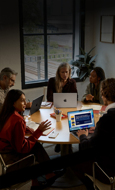 Cinco personas sentadas alrededor de una mesa de conferencias colaboran en sus MacBook, con sus iPhone boca abajo sobre la mesa