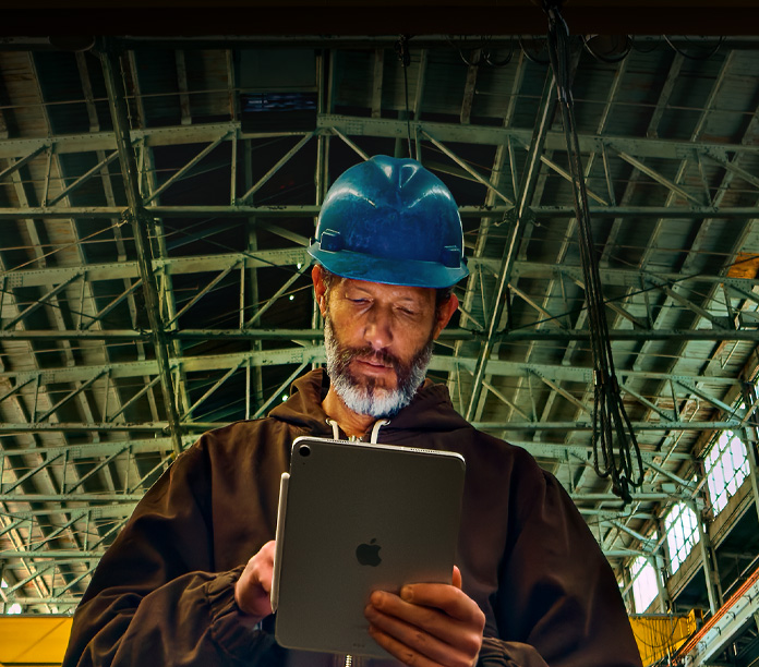 iPad, color plata, con un Apple Pencil adherido, sostenido por una persona con chaqueta café y casco azul, trabajando en un entorno industrial con vigas de metal