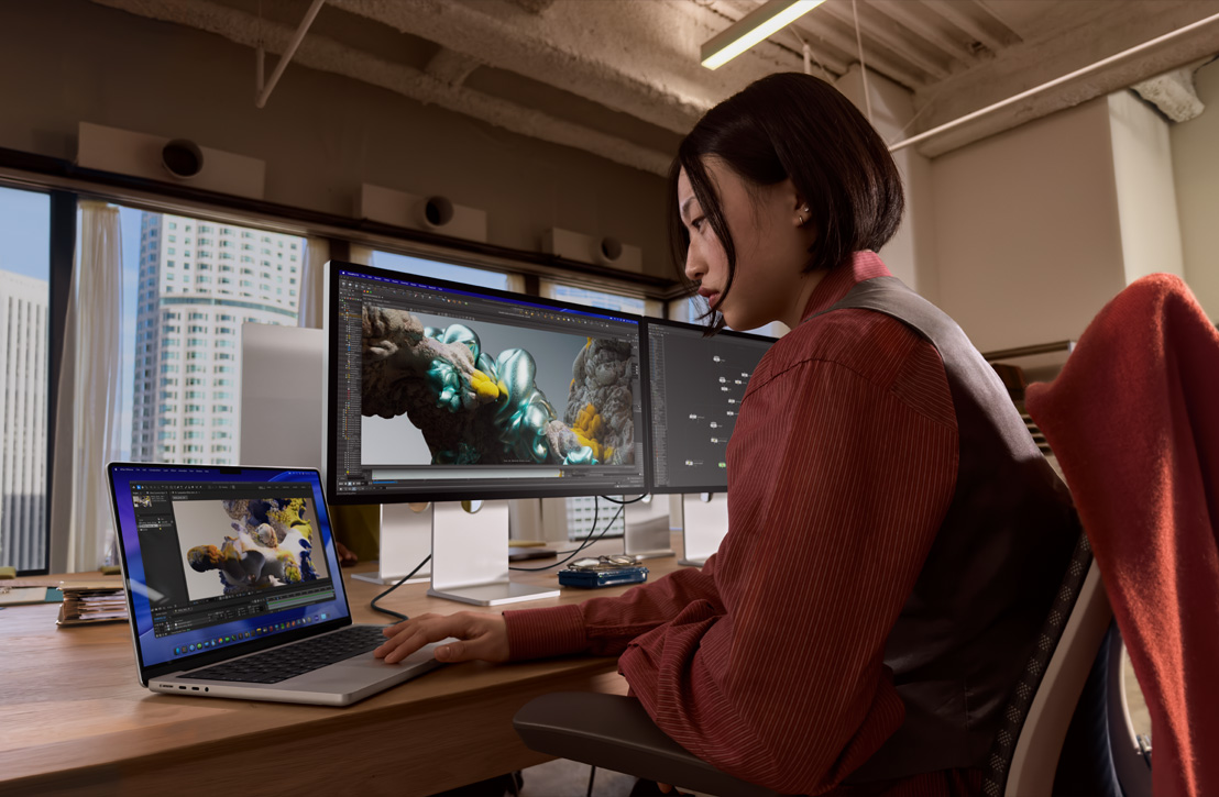 Una persona trabaja en un escritorio con una MacBook Pro conectada a un monitor externo, ambas pantallas muestran una renderización 3D, detrás se ven edificios a través de la ventana
