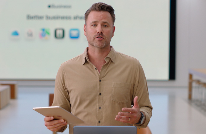 De presentator spreekt met een iPad in de hand, op de achtergrond is een groot scherm te zien waarop meerdere symbolen van zakelijke apps te zien zijn.