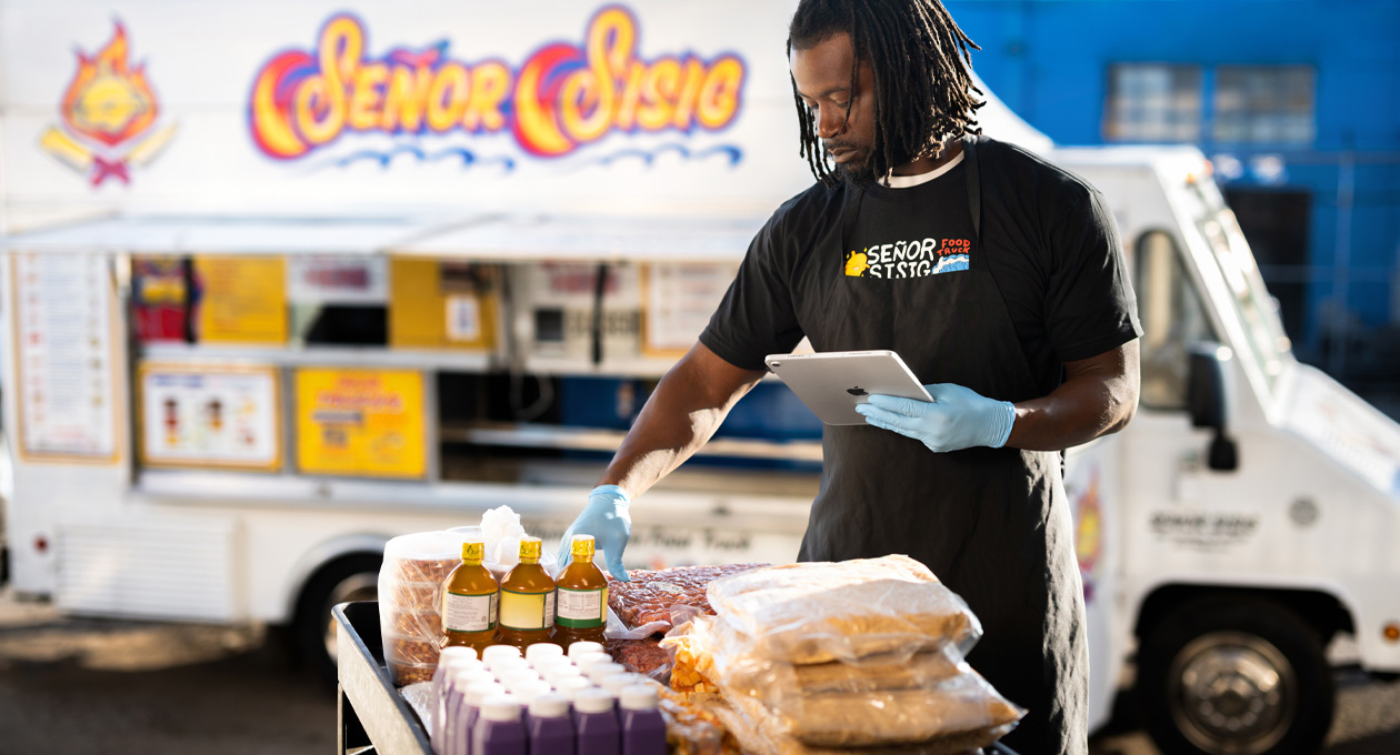 Una persona con una camiseta de la empresa y guantes revisa un iPad mientras prepara comida en una mesa, con un food truck de fondo