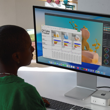 Una persona sentada trabaja en una hoja de cálculo de Excel titulada "Registro de ventas del cuarto trimestre" en una iMac y una Mac mini. Detrás de la hoja de cálculo de Excel hay una ventana de Pixelmator Pro y el fondo de macOS Tahoe.
