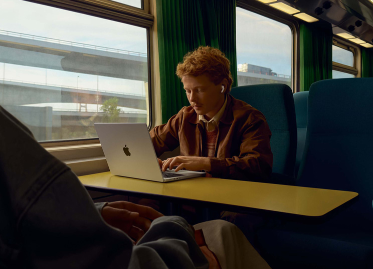 Person on a train at a window seat using AirPods and working on MacBook.