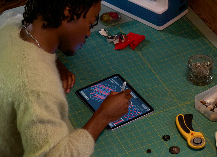 A person illustrating with Apple Pencil on iPad, seated at a green cutting mat with sewing supplies.