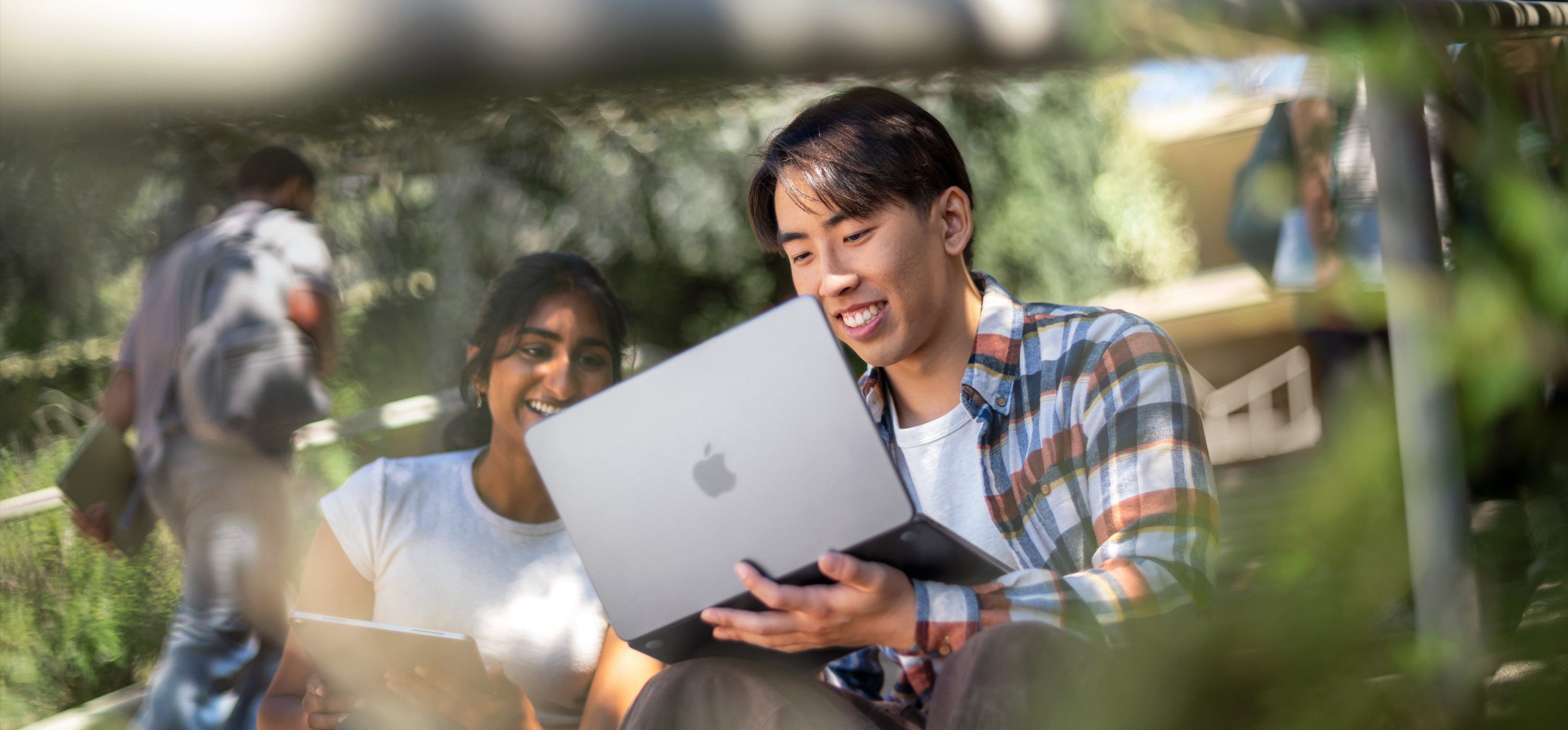 兩位學生面帶笑容在户外使用 MacBook，其中一位學生將 MacBook 放在大腿上，與另一位學生協作做功課