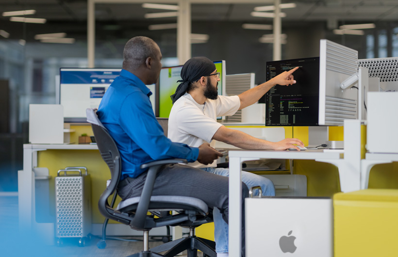 Two students sit in front of multiple iMac monitors, one pointing at a screen.