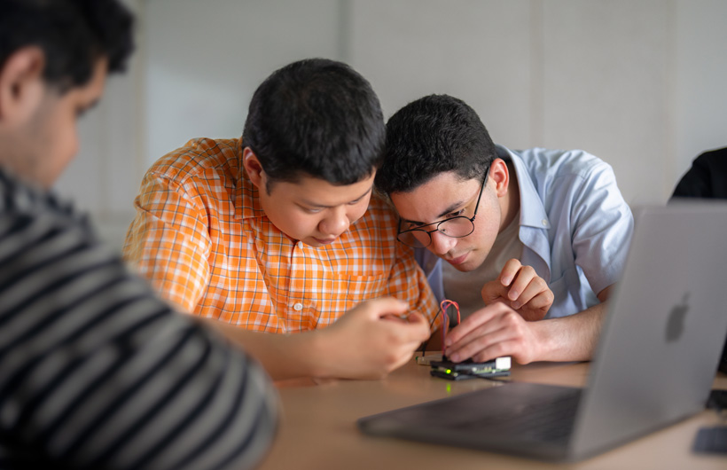 2 students closely examine a circuit board next to a MacBook, while a third looks on.