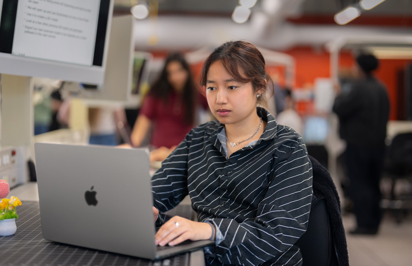 A student sitting at a desk in a modern classroom uses a MacBook.