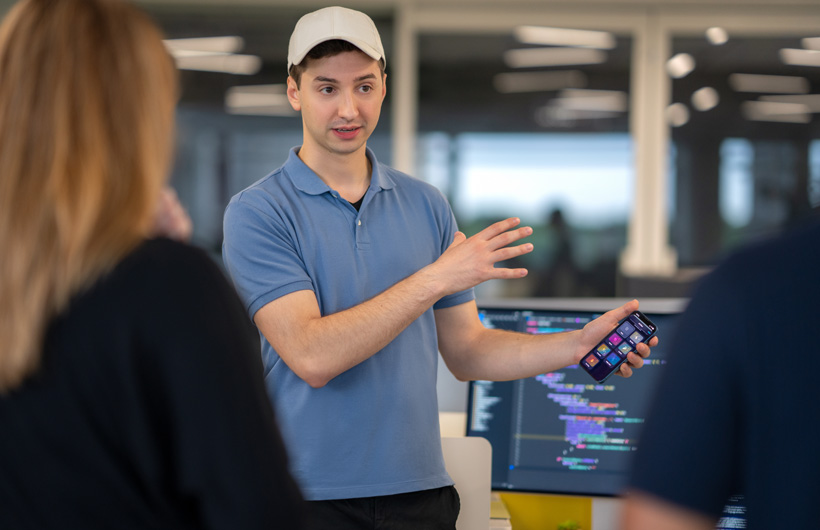 A student speaks to two people and gestures at an iPhone displaying a home screen of apps. Behind him, a monitor shows a code editor with colorful syntax-highlighted text.