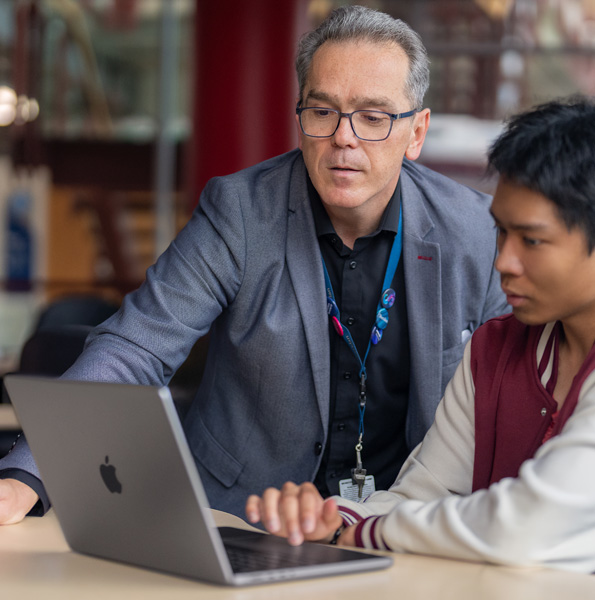 A man points to a laptop screen while a student sitting at a table in a classroom looks on.