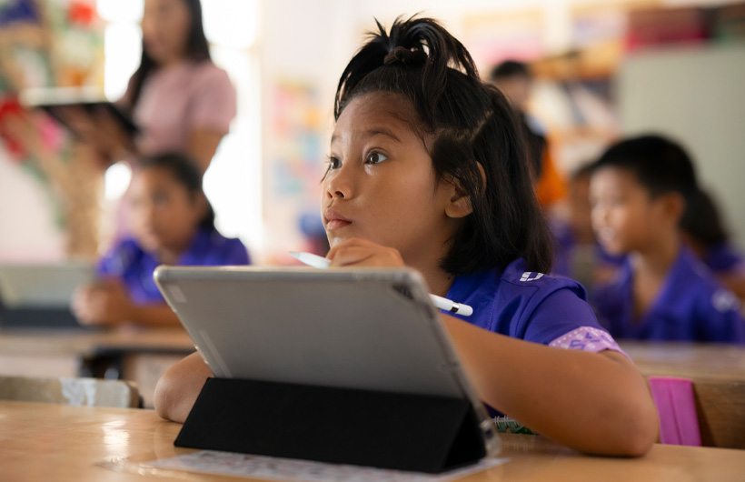 Ei ung jente sitter ved pulten sin og ser engasjert mot tavlen i klasserommet. Hun bruker en iPad og holder en Apple Pencil.