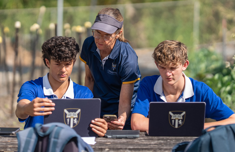 A teacher holds an iPhone and two students review information on their Mac while sitting on a bench in a garden.