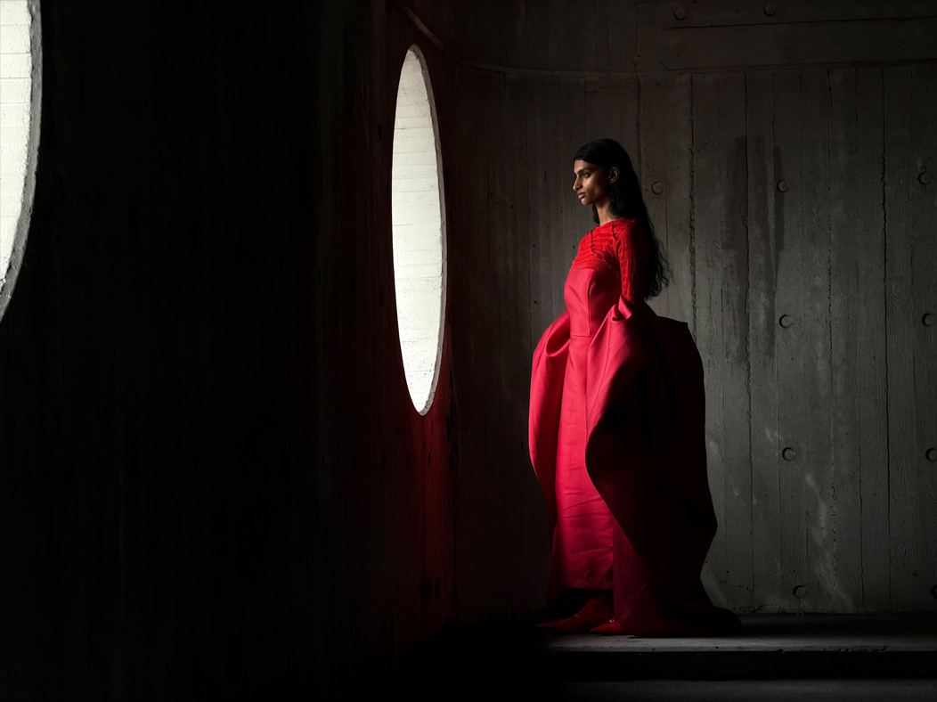 Une femme vêtue d’une longue robe rouge se fait photographier dans une pièce vide. L’app Appareil photo change de distance focale pour illustrer les capacités du système caméra pro, mettant en valeur: 100 mm