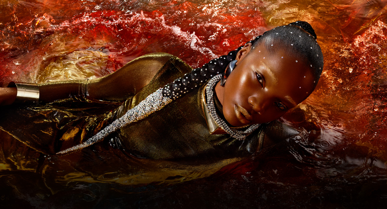 Ein Model in einem metallischen goldenen Kleid liegt auf der Seite in einem roten Wasserbecken, um die Helligkeit, den Kontrast und den grossen Farbraum des Studio Display XDR zu demonstrieren